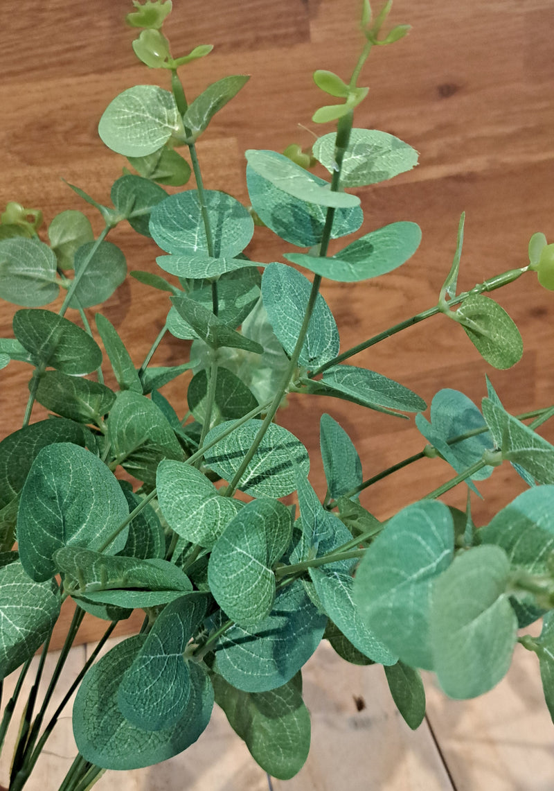 Artificial eucalyptus branch with green, round, textured leaves featuring prominent silver-gray veining, set against a warm brown wooden background.