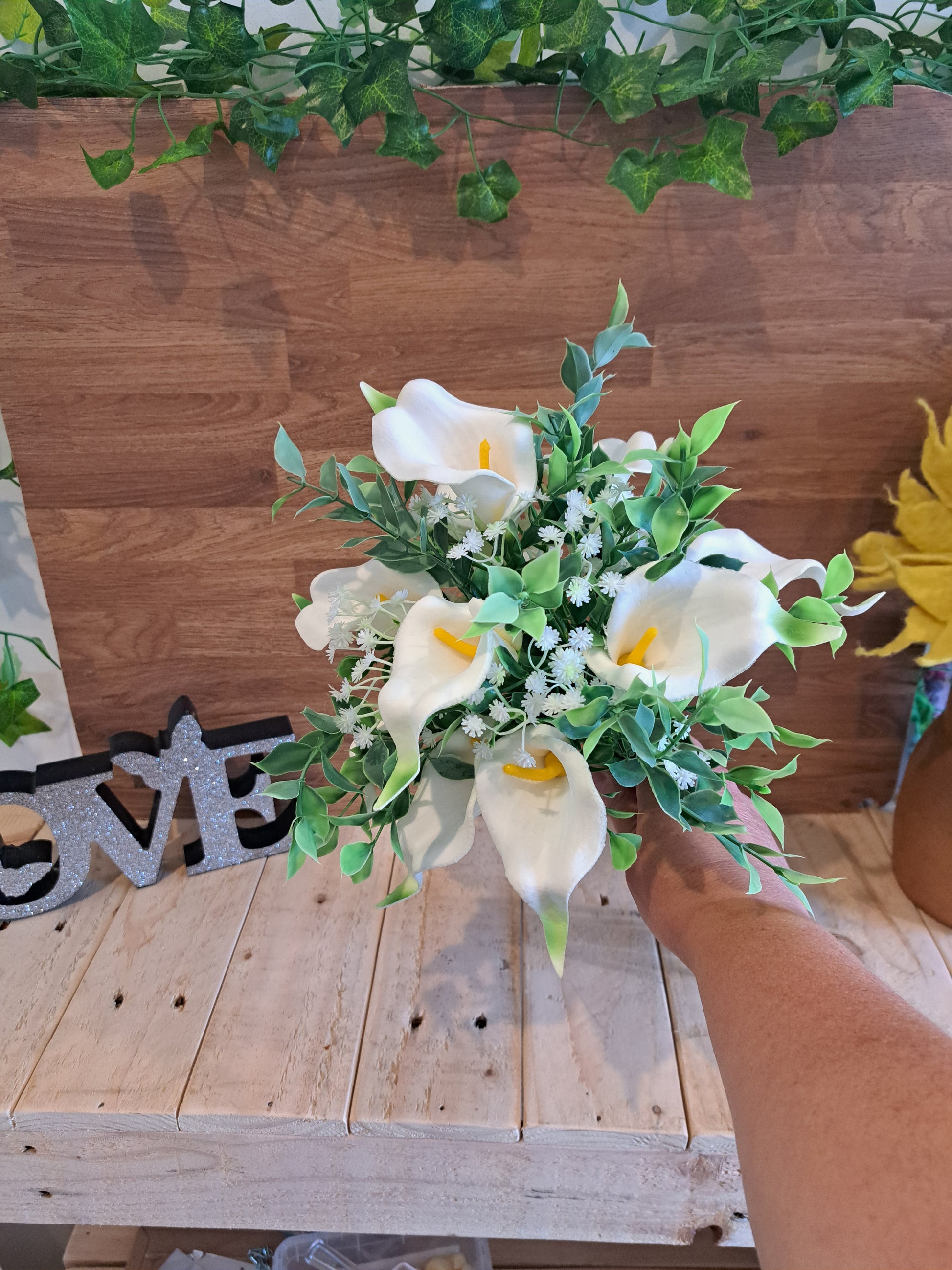 A bouquet of artificial white calla lilies and green leaves held by a person's hand in front of a wooden wall and bench.