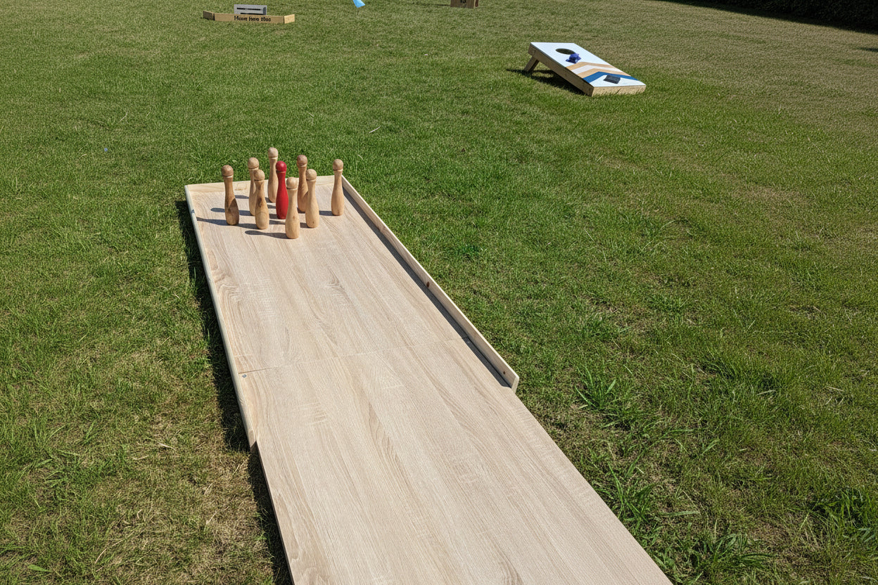 A wooden outdoor bowling alley game set on a grassy lawn, featuring a wooden alley with a cluster of wooden pins at the top. A cornhole board is visible in the background on the grass. Charity event, Southport