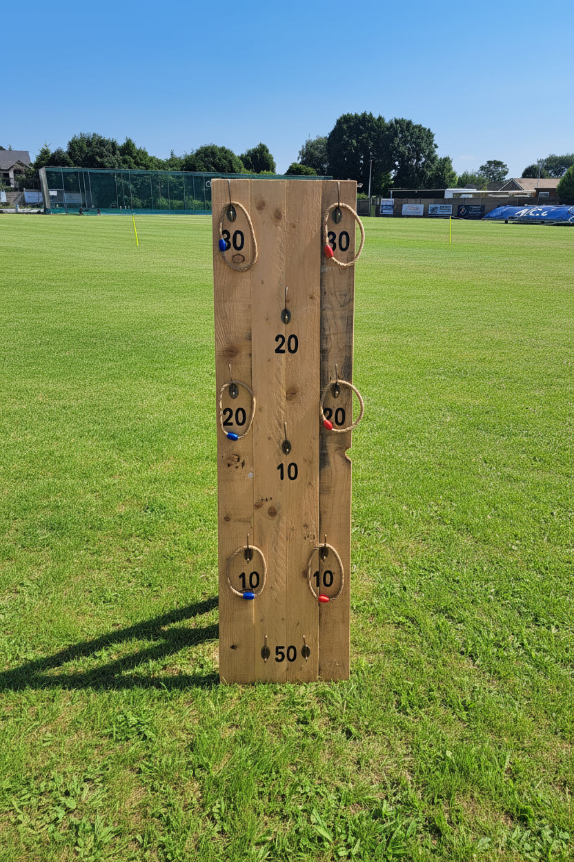 A wooden ring toss game board standing on a green grass field under a blue sky, with trees and buildings in the background. Charity event, Southport