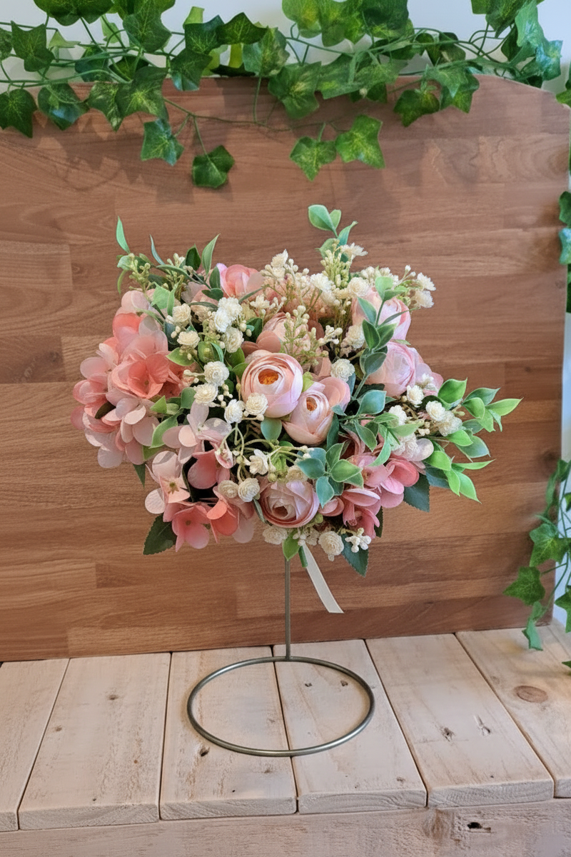 A faux floral arrangement in shades of pink, peach, cream, and green, displayed on a metal stand against a wooden wall and light wood tabletop.