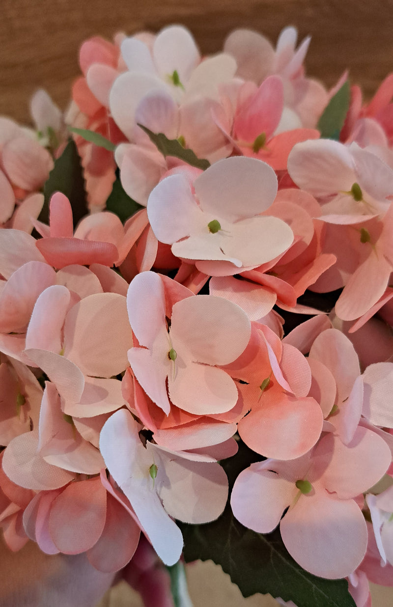 A close-up of a bouquet of artificial pink hydrangea flowers with green leaves, set against a warm brown wooden background.