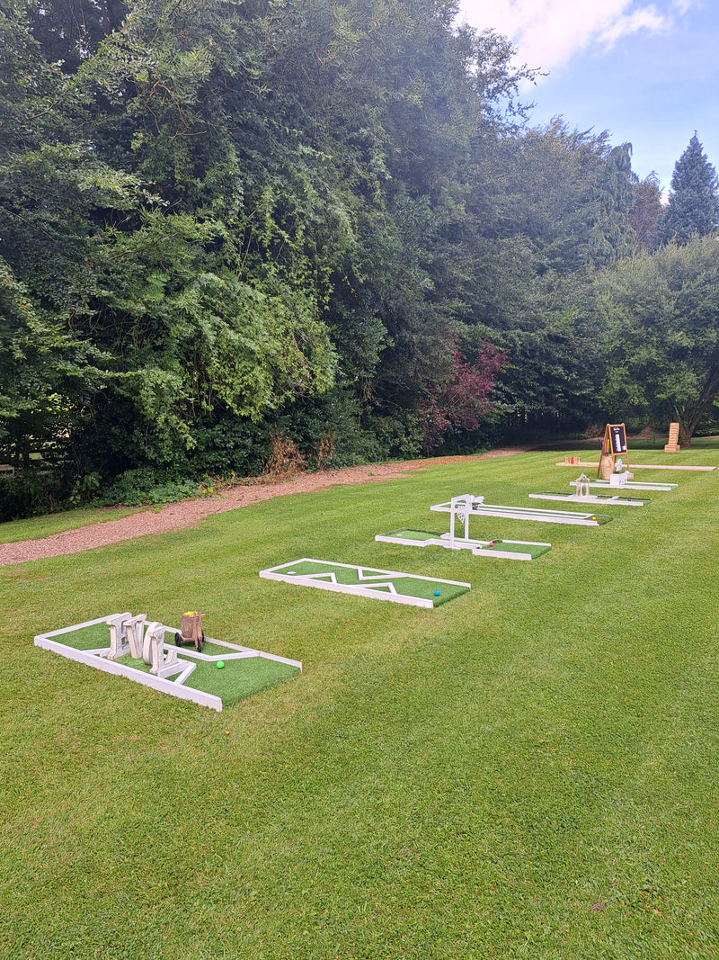 Outdoor mini golf course setup on a grassy lawn with trees in the background. Several white-framed golf holes are arranged in a row. Mitton Hall, Clitheroe