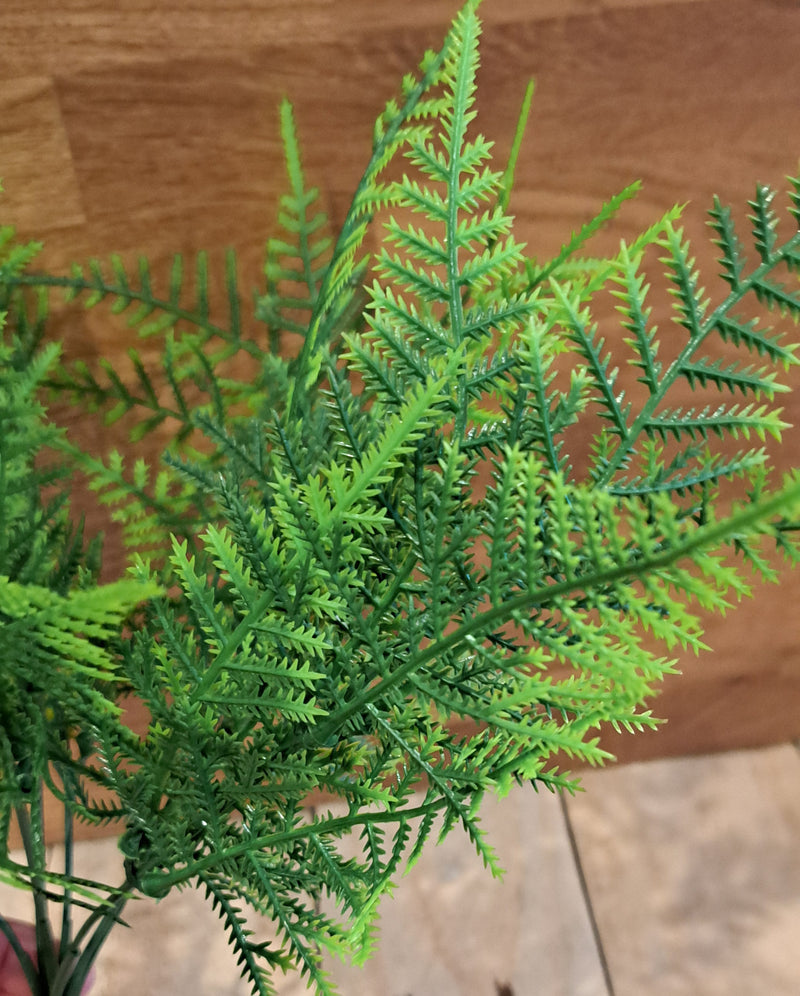 A close-up of a green artificial fern plant with detailed, feathery fronds, set against a background of a wooden wall and tiled floor.