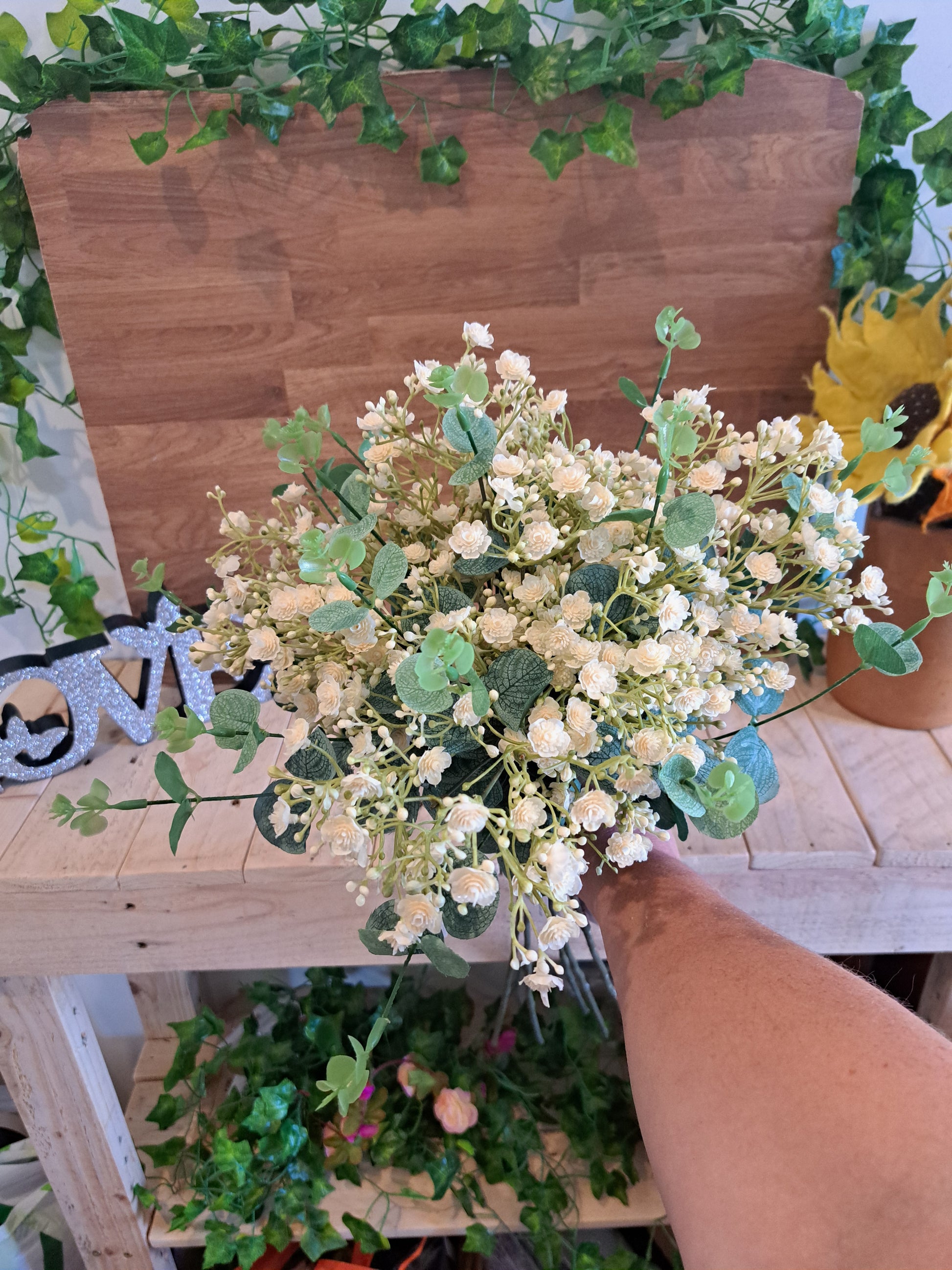 An artificial floral bouquet with small cream-white flowers and green leaves, held by a person's hand in front of a wooden shelf decorated with greenery.