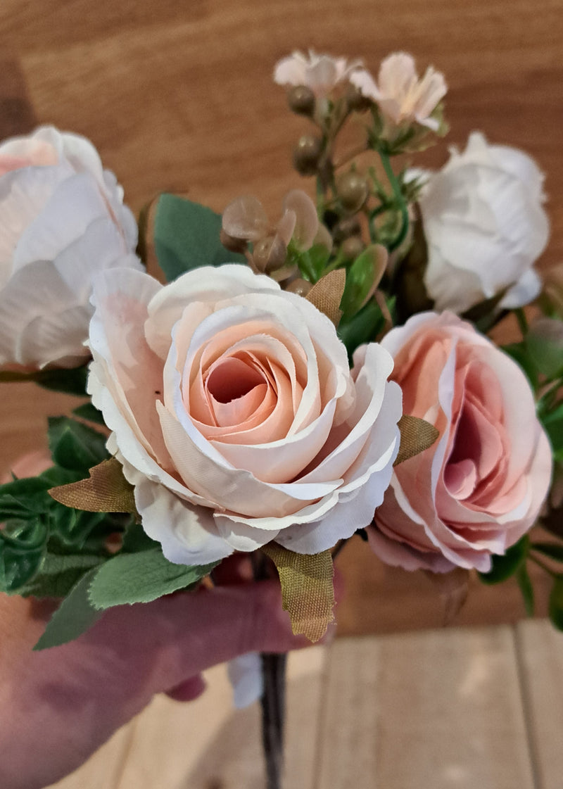 A close-up of a faux floral arrangement featuring pale blush pink and white roses with green leaves, held by a hand against a wooden background.