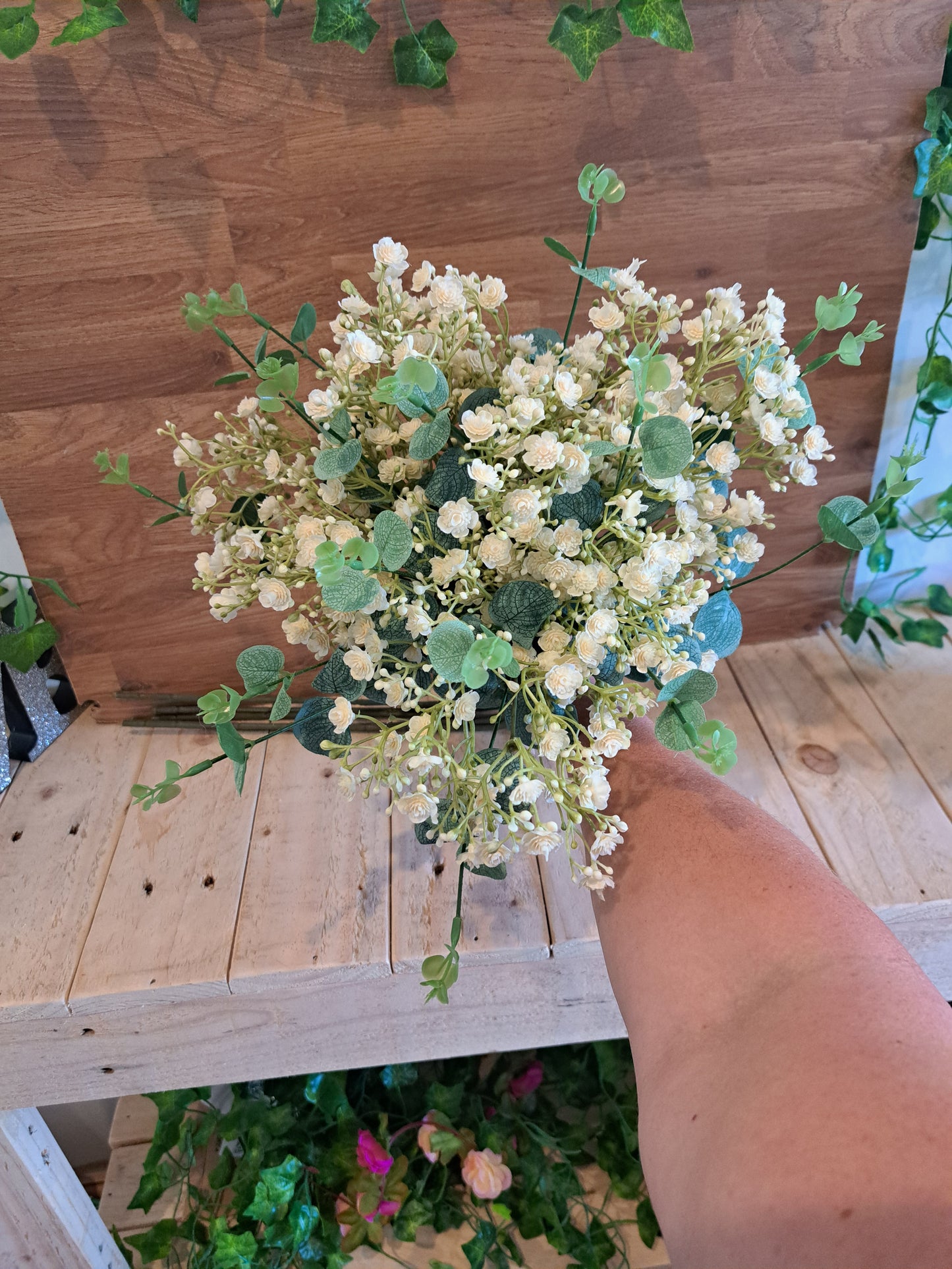 A bouquet of artificial ivory gypsophila flowers with green eucalyptus leaves, held by a person's arm against a rustic wooden shelf background.