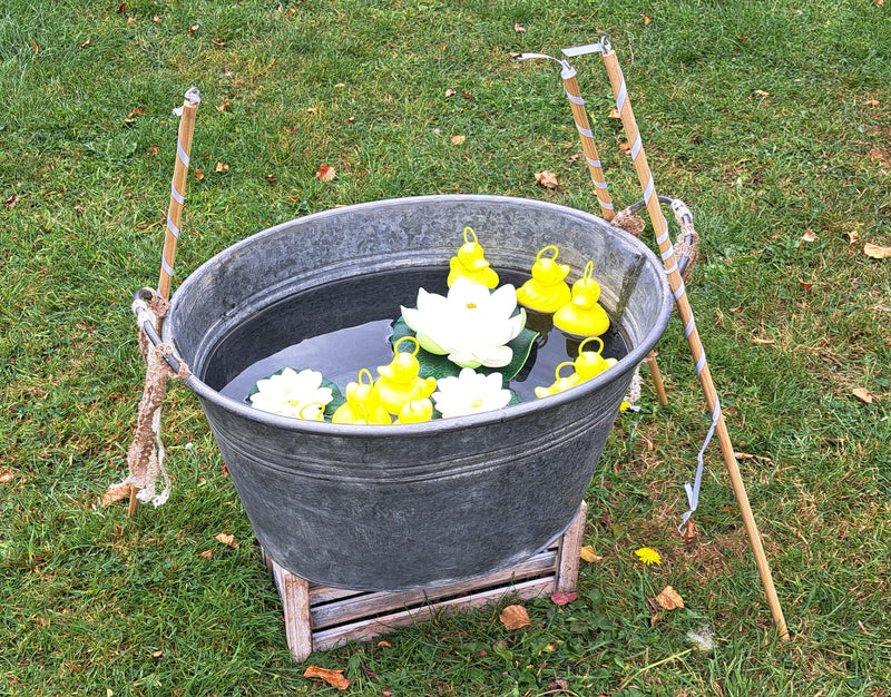 A galvanized metal tub filled with water and floating white lotus flowers and yellow ducks, set on a small wooden stand outdoors on a grassy lawn for a wedding.