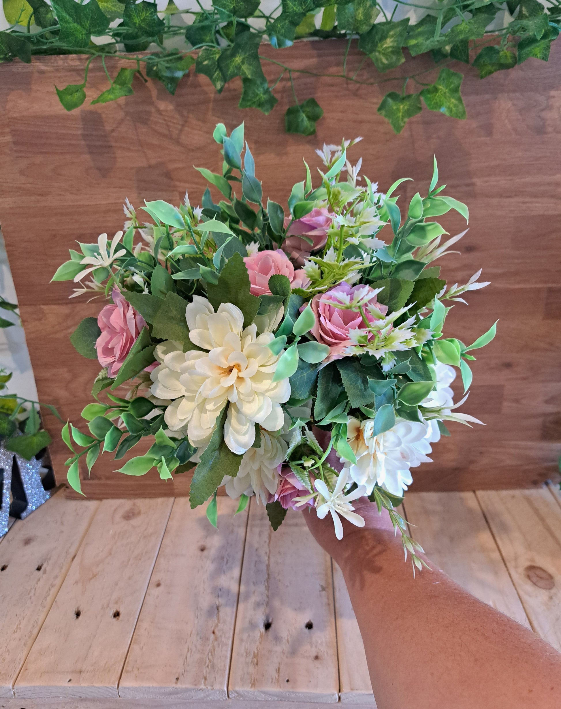 A hand holding a bouquet of artificial flowers, including cream dahlia-like blooms, pink roses, and green foliage, set against a wooden wall and floor.