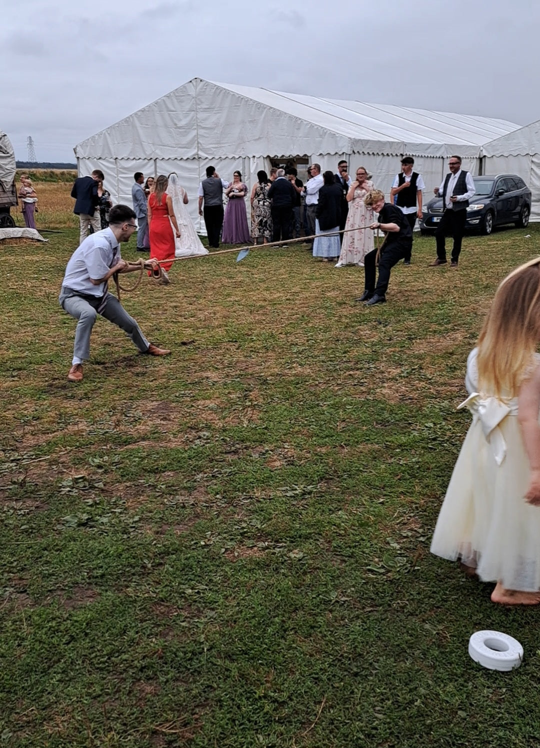 An outdoor wedding on a grassy field with a large white marquee tent in the background. Several guests are standing and mingling near the tent, while two men in the foreground are having a game of tug of war, and a young girl in a white dress stands in the right foreground.