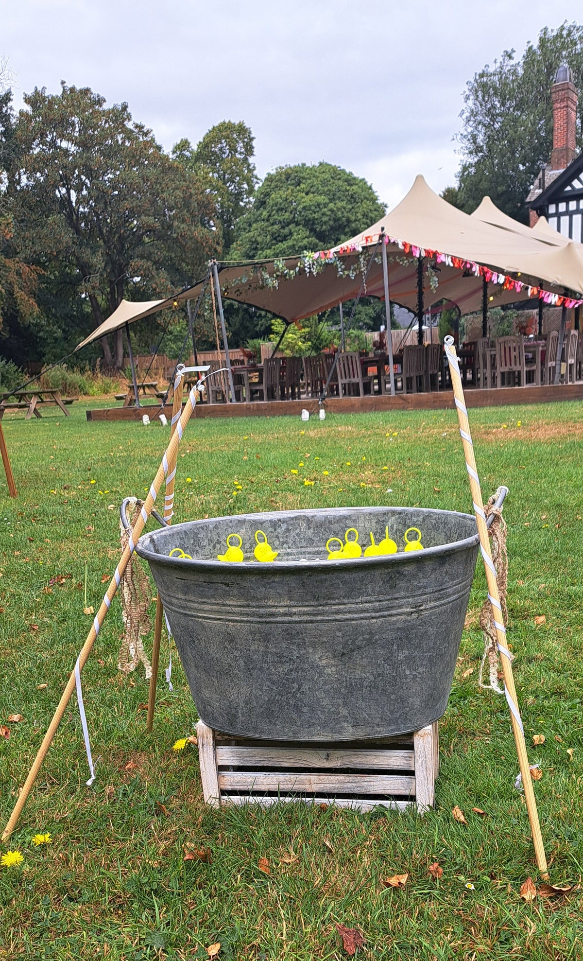 A large galvanized metal tub elevated on a wooden stand outdoors on a grassy field, with poles and small yellow ducks inside the tub. A large pavilion and trees are visible in the background under an overcast sky.