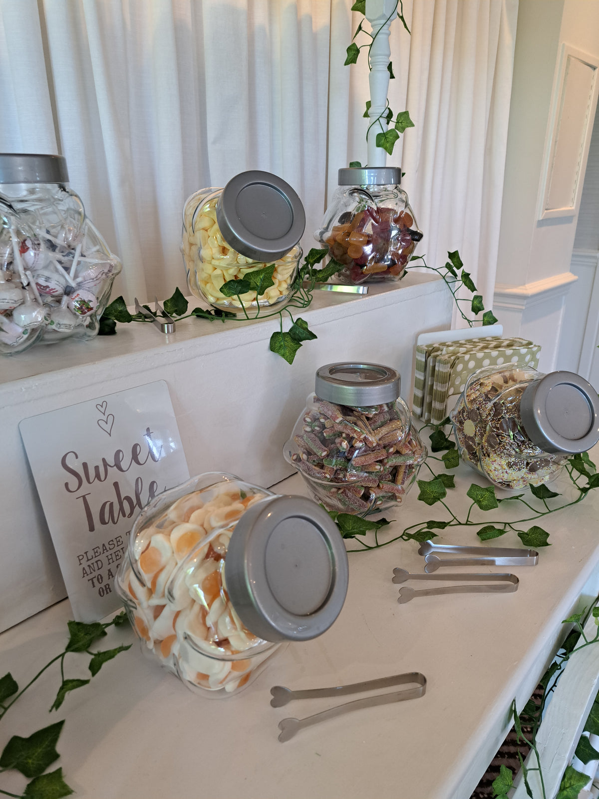 A sweet table candy display with several clear glass jars filled with assorted candies and lollipops, arranged on a white sweets cart with silver metal tongs, green ivy vines, and a white sign that reads 'Sweet Table'. 