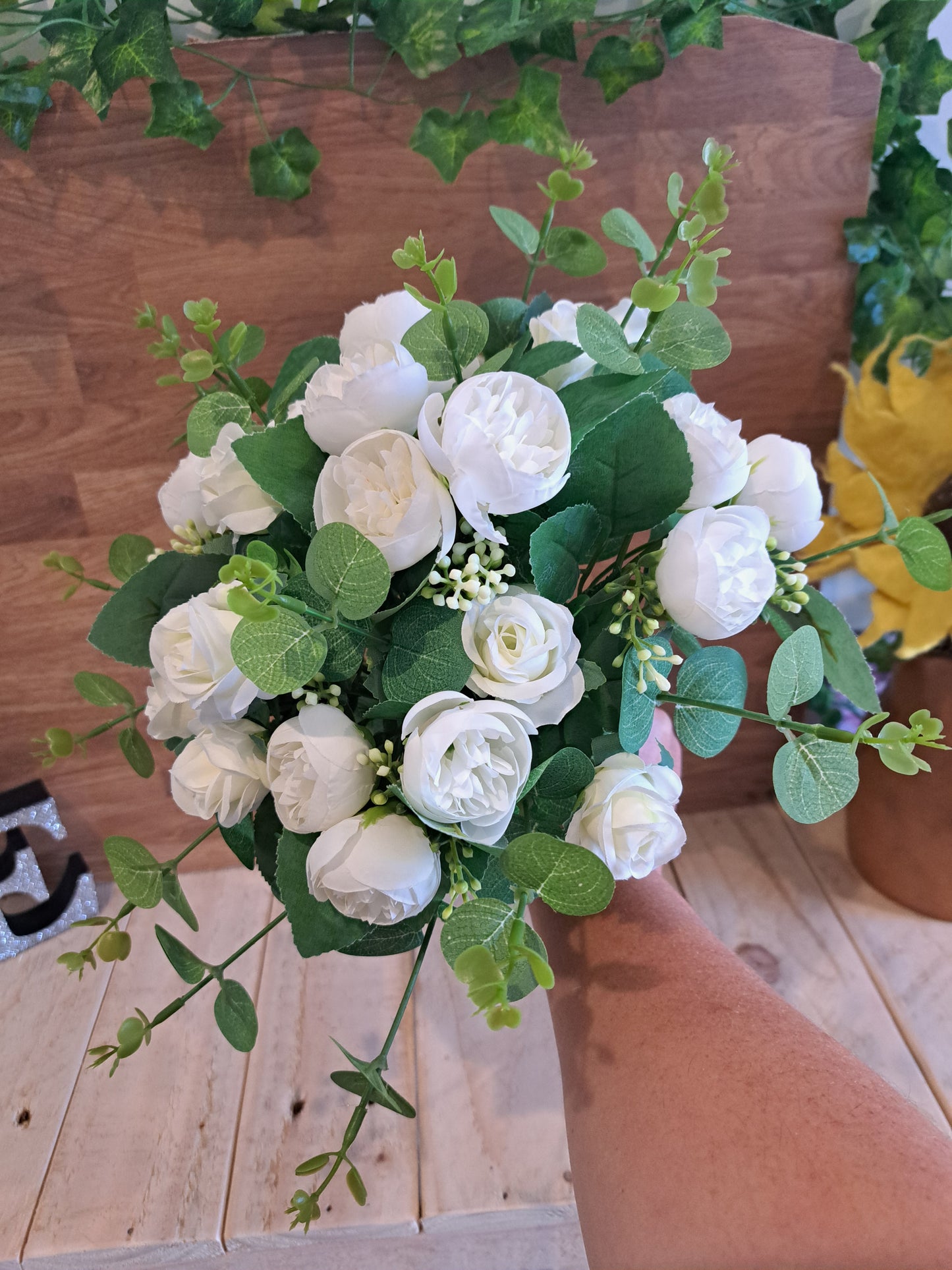A hand holding a bouquet of white faux roses and green eucalyptus leaves in front of a wooden wall and tabletop.