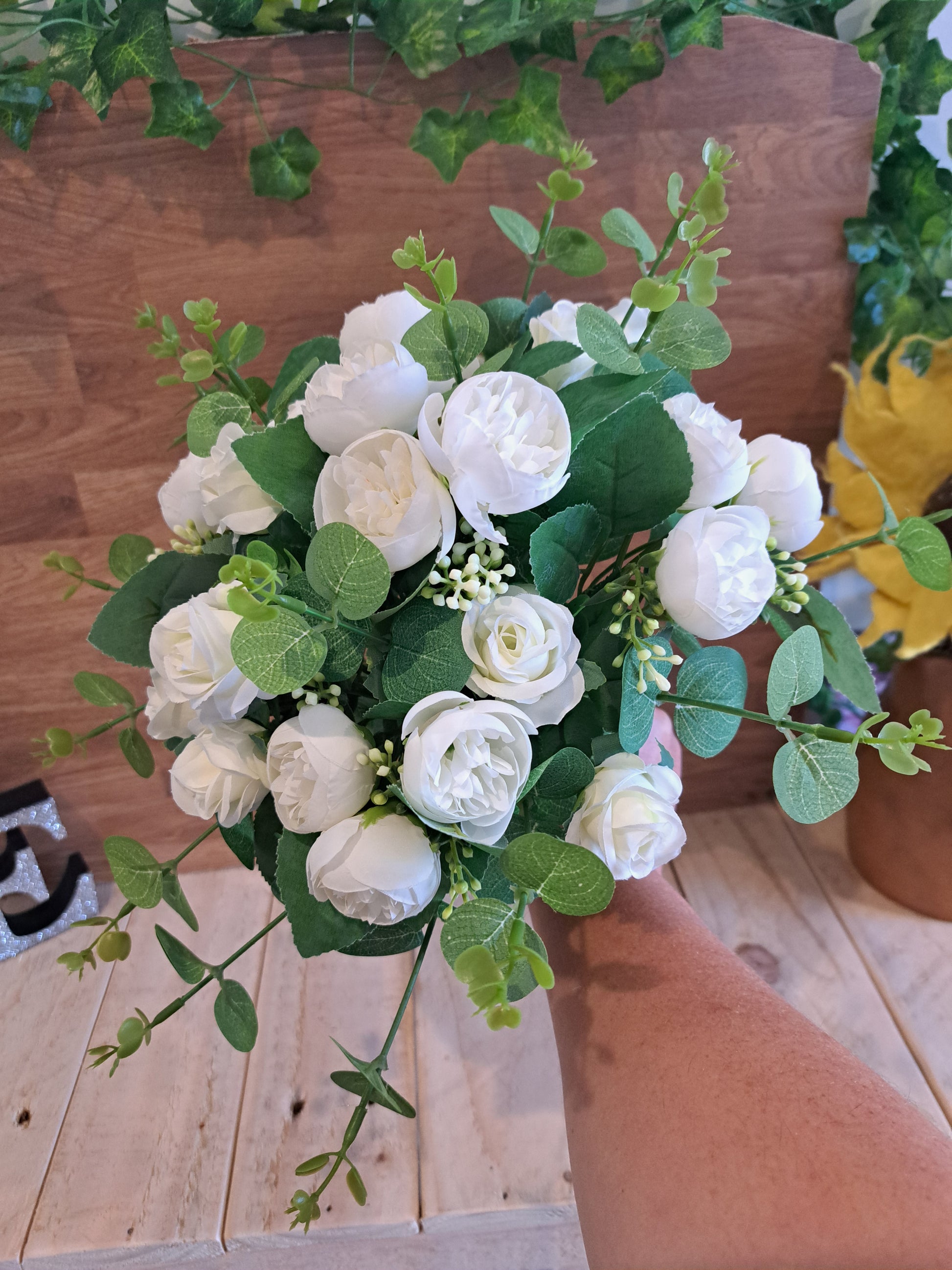 A hand holding a bouquet of white faux roses and green eucalyptus leaves in front of a wooden wall and tabletop.