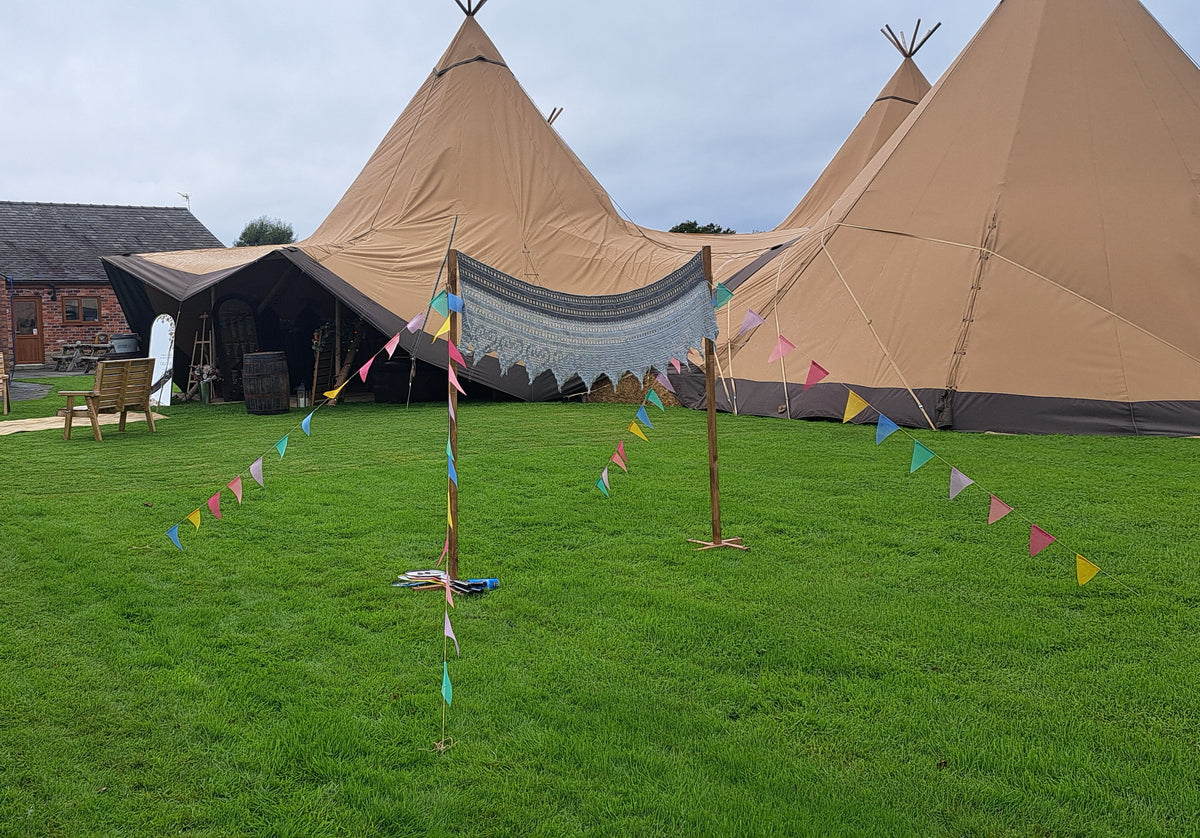 Large tan wedding tipi tents set on a green lawn, a boho badminton net, with colourful bunting strung between wooden poles in the foreground. A brick building is visible on the left under an overcast sky. Parr Hall Farm, Eccleston 