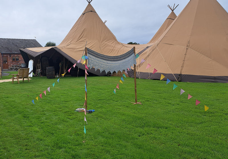 Large tan wedding tipi tents set on a green lawn, a boho badminton net, with colourful bunting strung between wooden poles in the foreground. A brick building is visible on the left under an overcast sky. Parr Hall Farm, Eccleston 