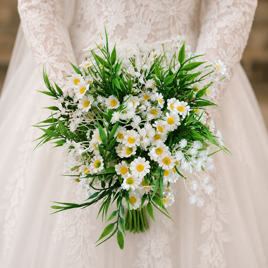 A bridal bouquet of white daisies and baby's breath with green leaves held by a person wearing a white lace wedding dress.