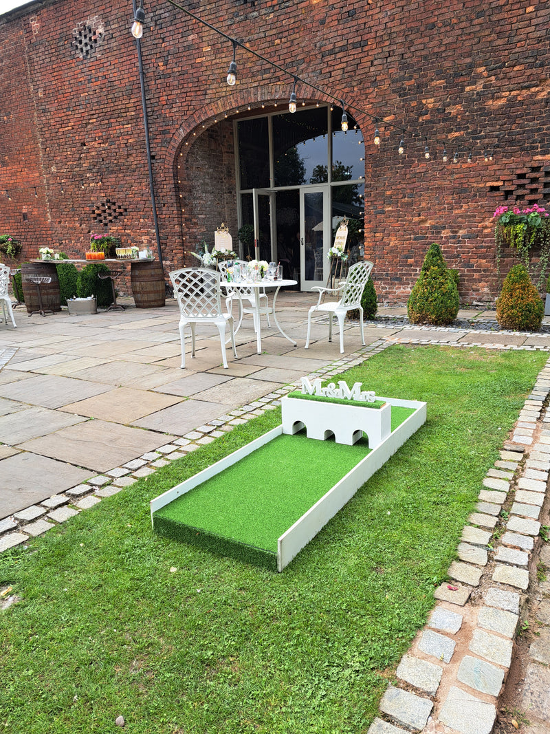 Outdoor setting with a brick building, green lawn, and white chairs with a wedding mini golf hole in the for ground. 