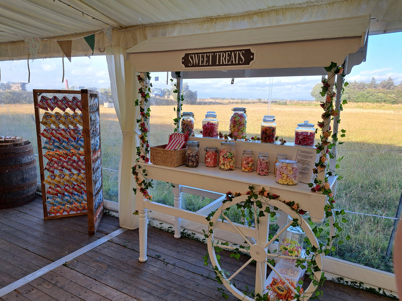 A white wooden 'Sweet Treats' candy cart decorated with green ivy garlands, filled with jars of colourful candies and sweets, and a large rustic crisp wall filled with crisps, set up in a marque for a wedding. Private garden, Southport.