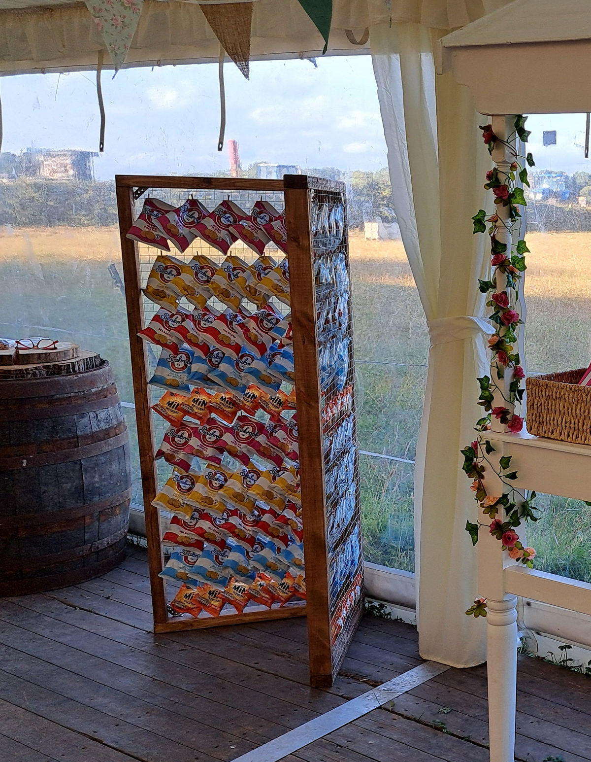 A wooden folding crisp wall display rack filled with colourful packaged crisp bags, set on a wooden deck inside a tent with a field view in the background.