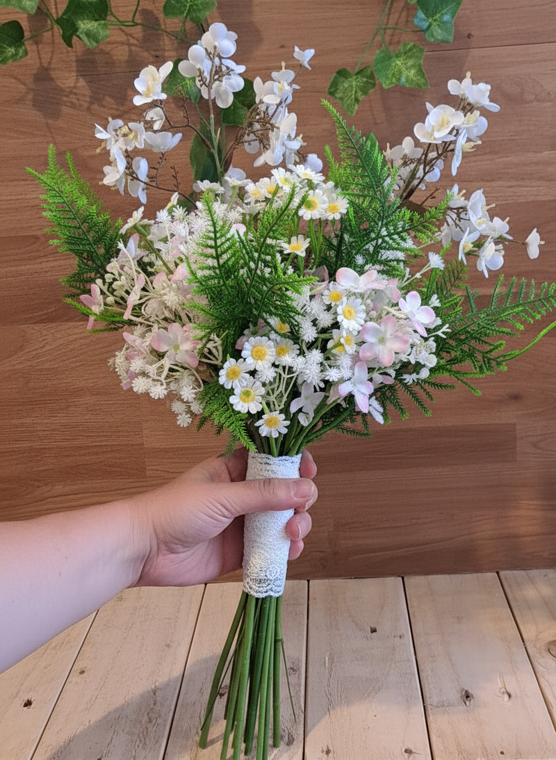 A hand holding a bouquet of artificial white and pale pink flowers with green fern-like foliage, wrapped with a white lace ribbon at the stem, set against a wooden plank background.