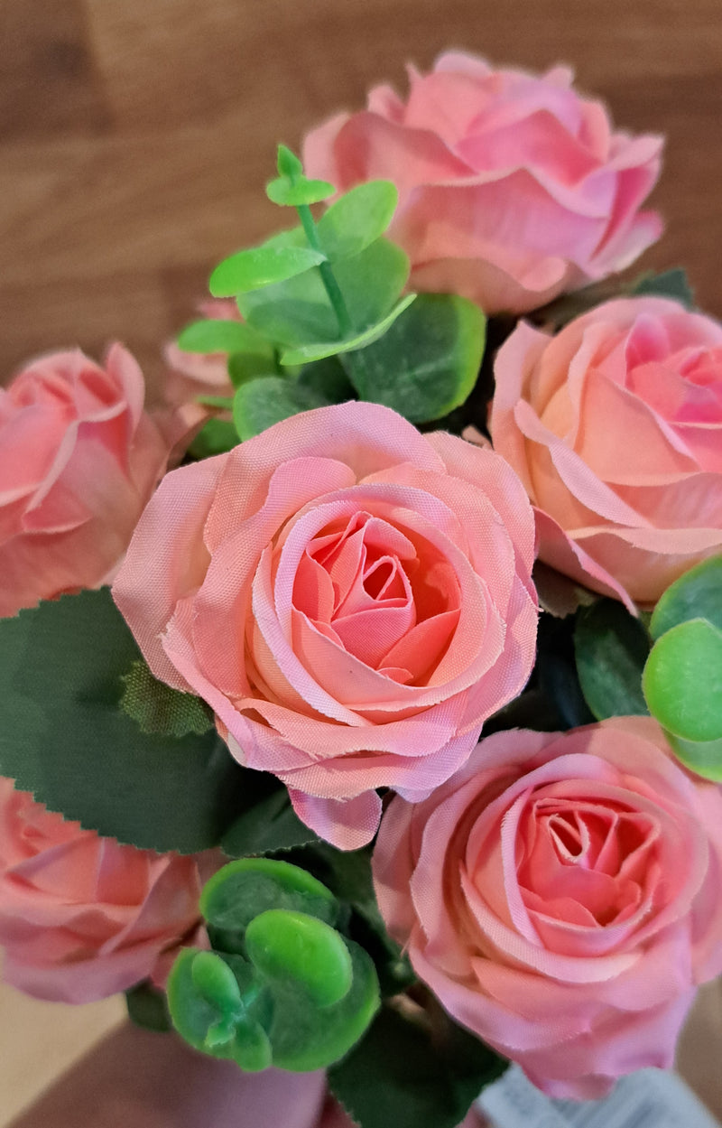 Pink faux rose artificial flower bouquet with green leaves, held by a hand against a wooden background.