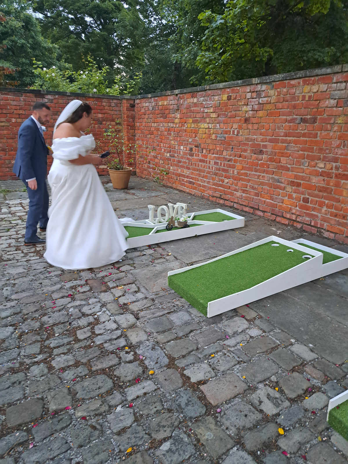 A bride in a white wedding dress and groom in a blue suit playing mini golf on a cobblestone courtyard, with a brick wall and trees in the background. Meols Hall
