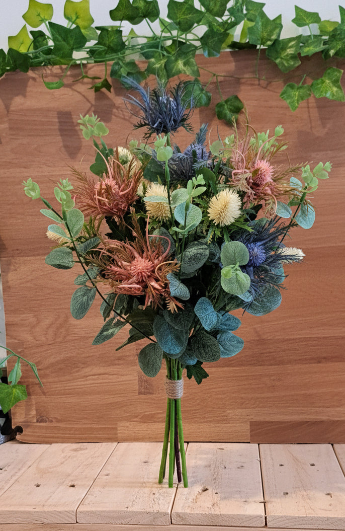 A rustic, multi-colored artificial flower bouquet with green eucalyptus leaves, blue thistle-like flowers, and dried orange and pink accents, set against a wooden shelf and ivy backdrop.