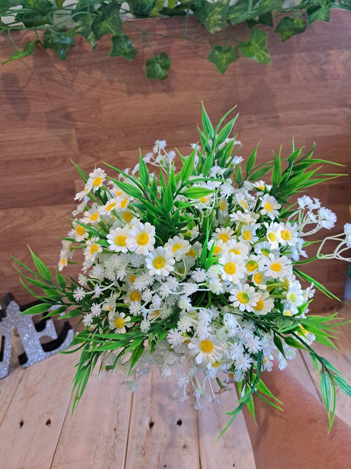 An artificial daisy bouquet with green leaves and white baby's breath, held by a hand against a rustic wooden plank background.