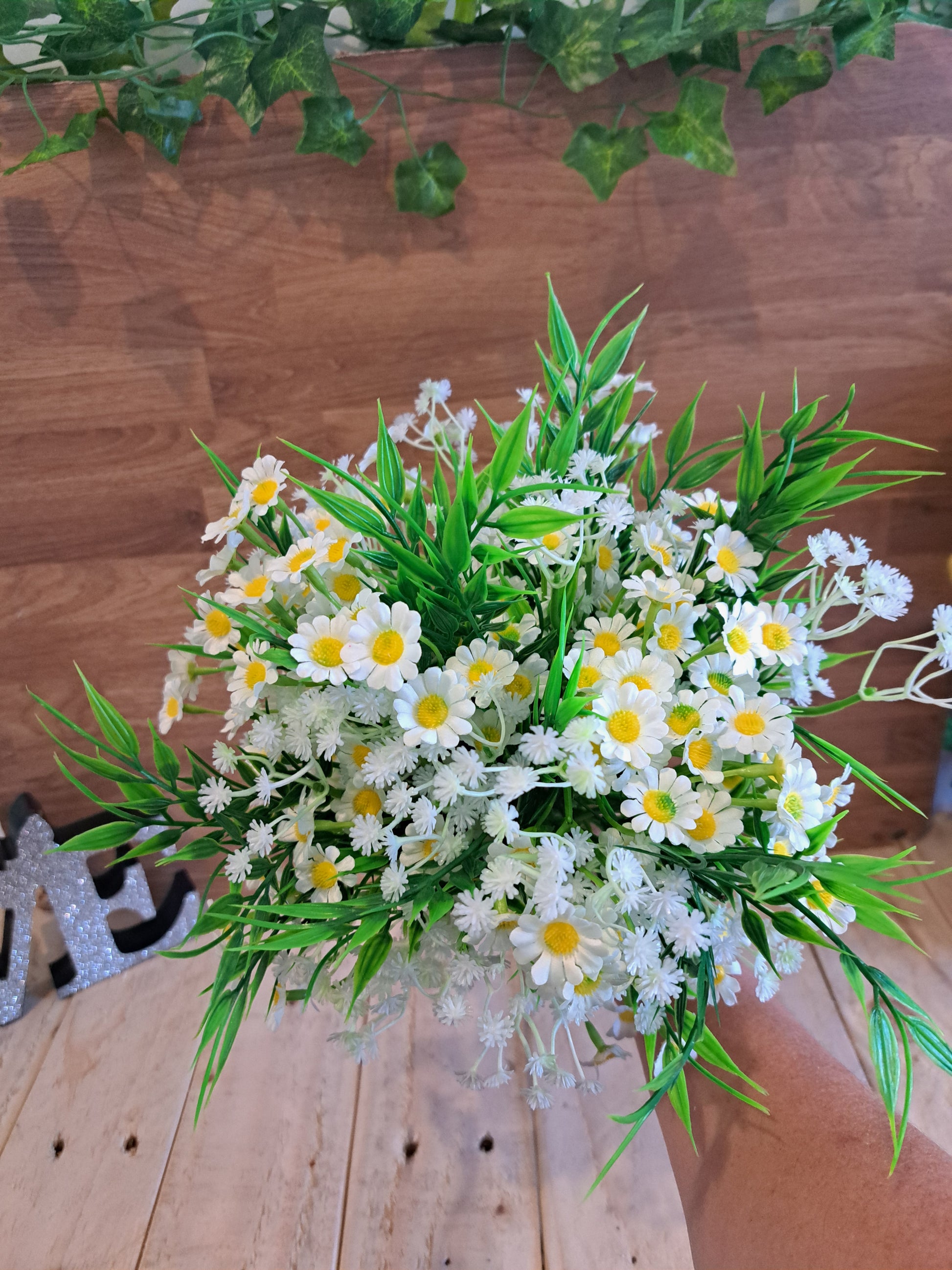 An artificial daisy bouquet with green leaves and white baby's breath, held by a hand against a rustic wooden plank background.