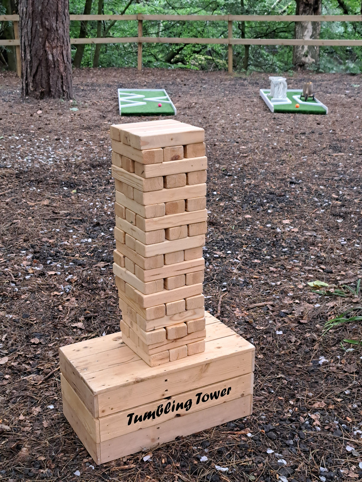 A giant wooden tumbling tower game set in a wedding woodland setting, mini golf holes are visible in the background near trees and a fence. Orrell Hill Woods, Ince Blundell