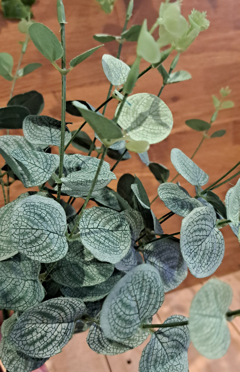 Artificial eucalyptus branch with dusty green, round, textured leaves featuring prominent silver-gray veining, set against a warm brown wooden background.