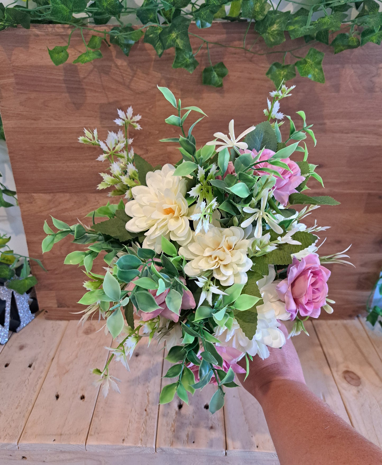 A hand holding a bouquet of artificial flowers, including pink and ivory roses with green leaves, set against a rustic wooden plank background.