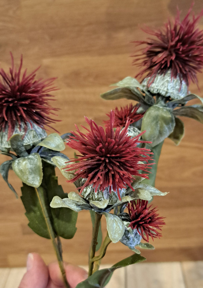 A bouquet of artificial red spiky thistle flowers with green leaves, held by a hand against a wooden background.