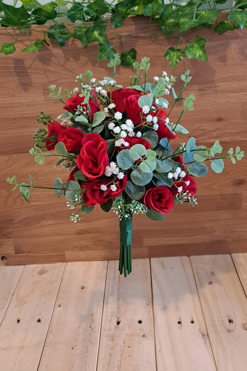 A bouquet of red roses, green eucalyptus leaves, and small white baby's breath flowers in front of a wooden wall with green ivy.
