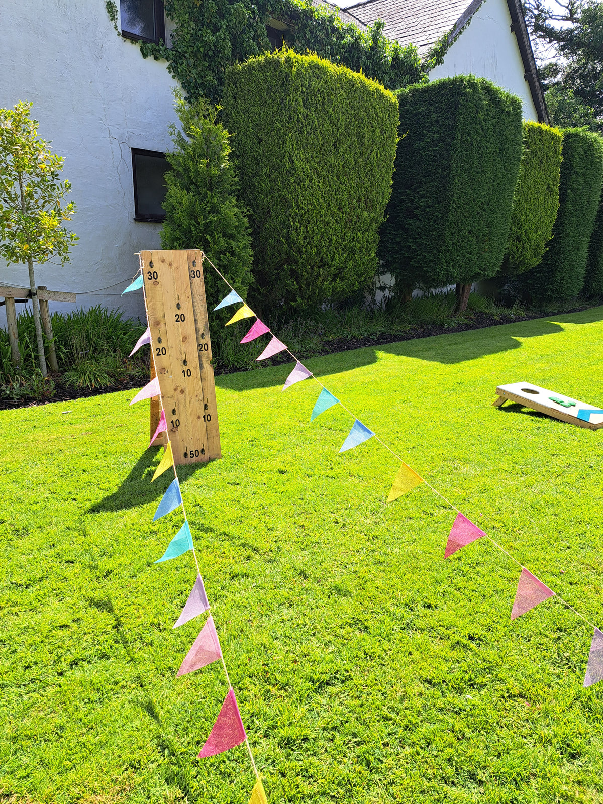 Outdoor wedding games area with a wooden ring toss game on a string of colourful bunting, set on a green lawn with hedge hedges and a house in the background. The River Barn, Poulton-le-Fylde