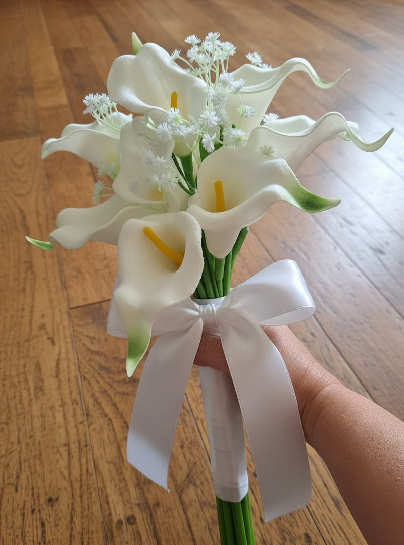 A hand holding a bouquet of artificial white calla lilies with green stems and small white filler flowers, photographed indoors against a wooden floor.