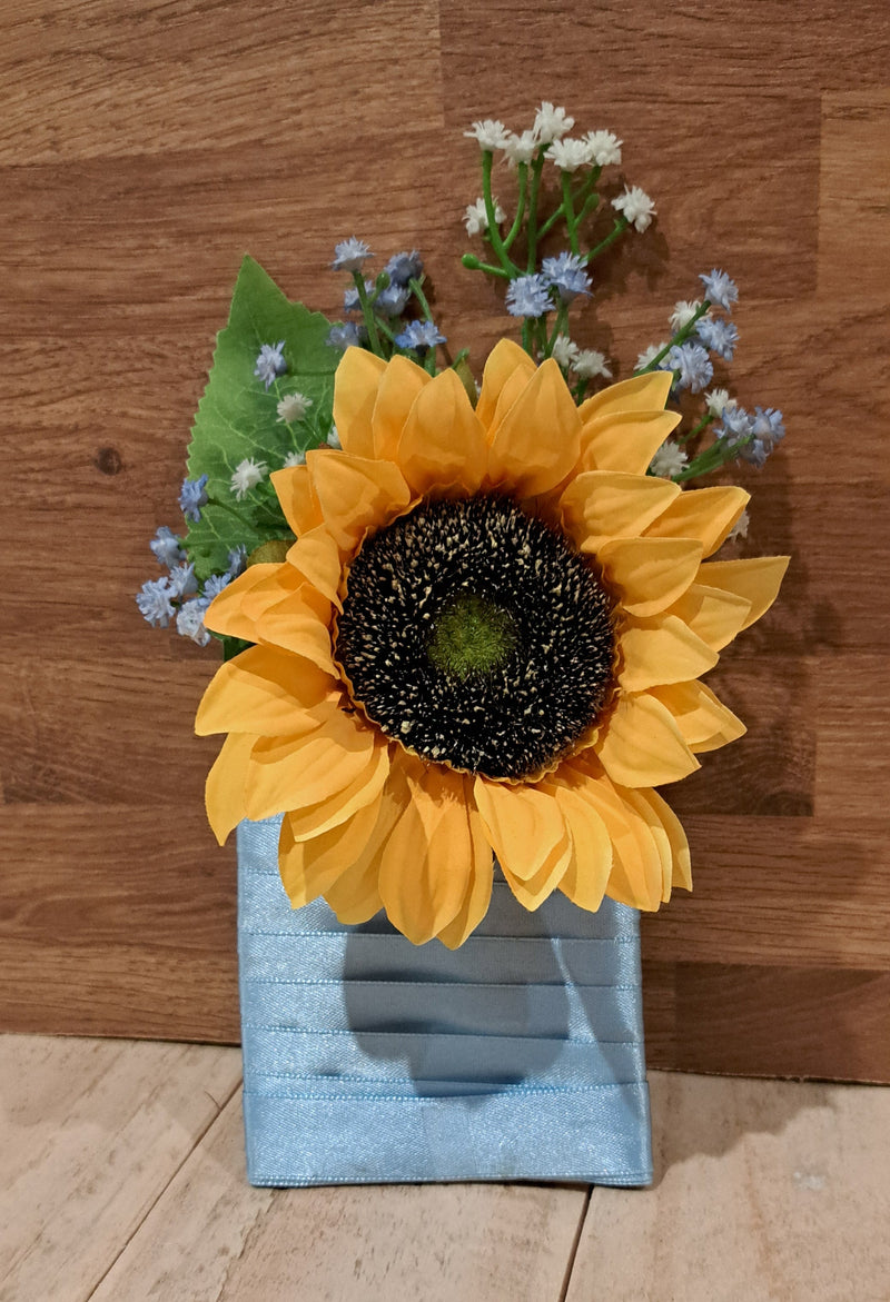 A single artificial sunflower with bright yellow petals, a dark brown to black center, green leaves, and small white and blue flowers, arranged in a light blue grooms pocket flower button hole against a wooden background.