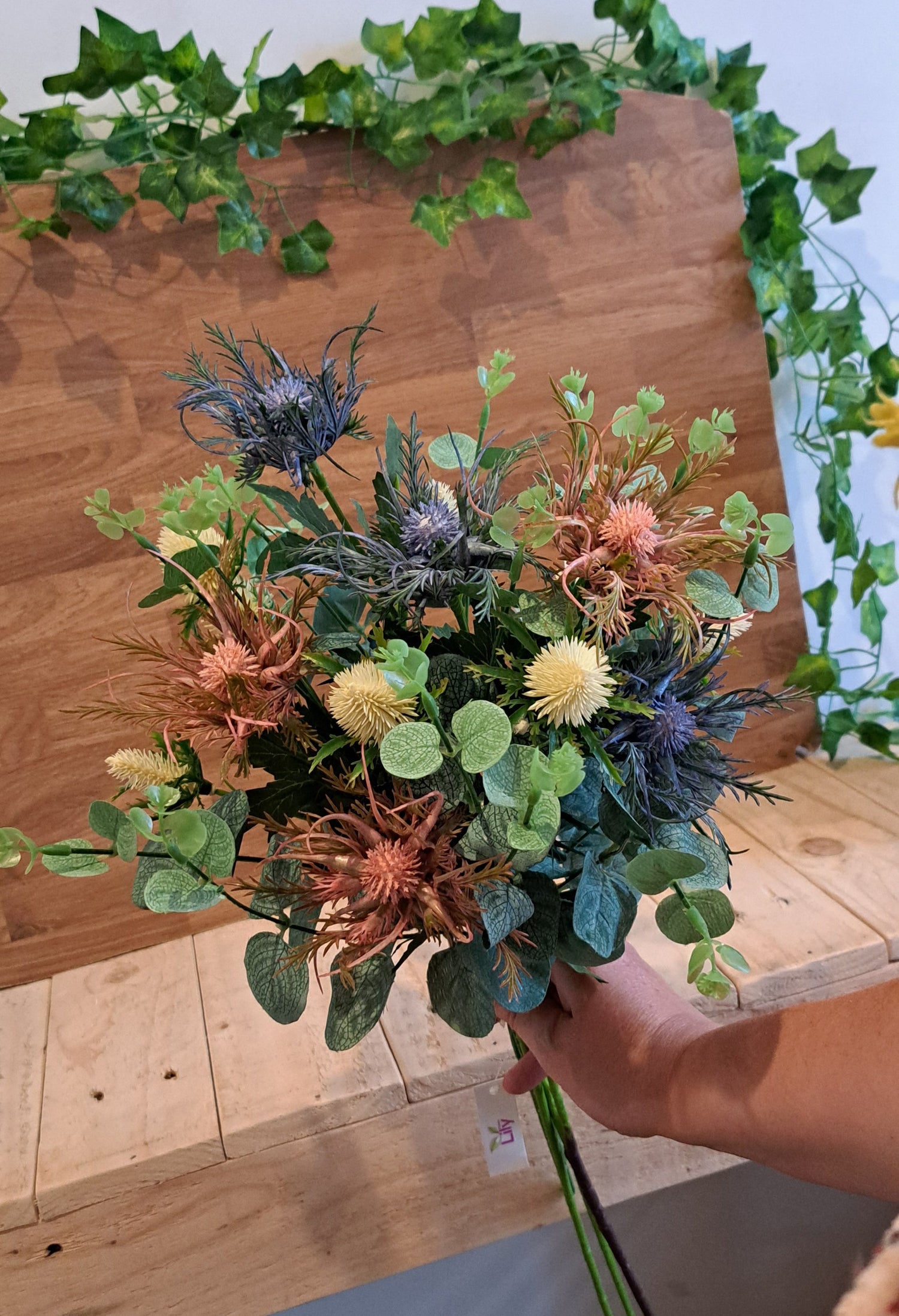 A hand holding a rustic dried flower bouquet with green eucalyptus leaves, blue thistles, and orange-brown wildflowers, set against a wooden display board with green ivy.