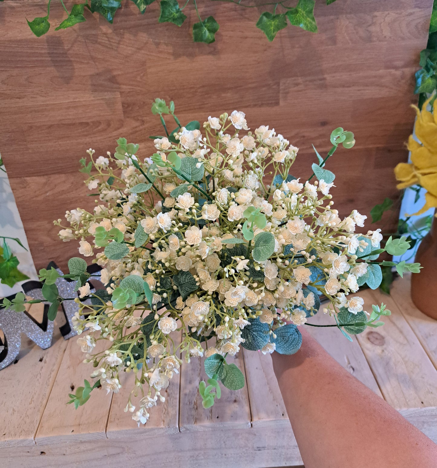An artificial floral bouquet with small ivory flowers and green leaves, held by a person's arm against a wooden wall and tabletop background.