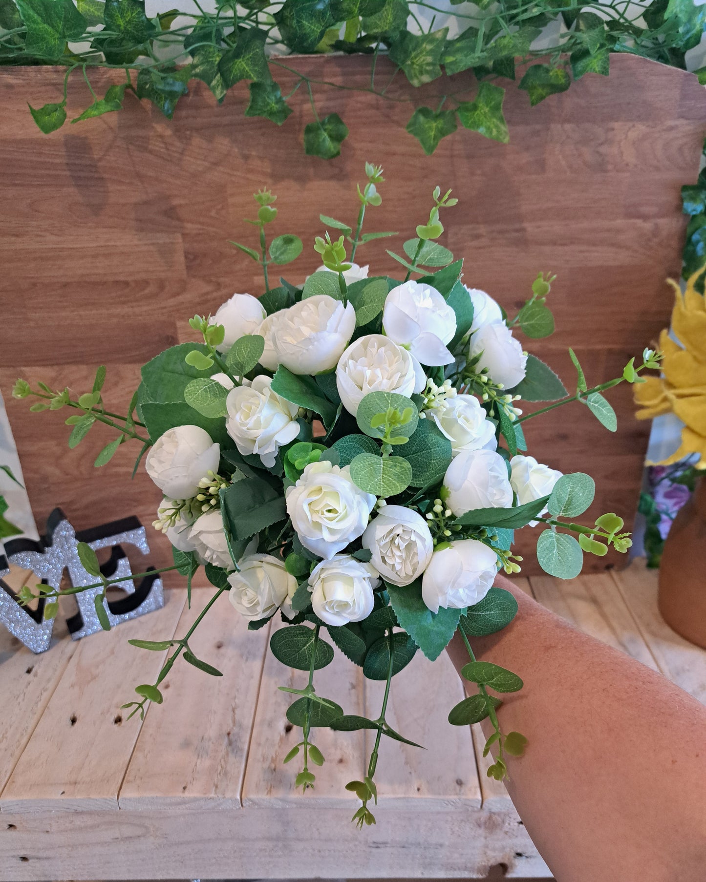 A bouquet of white faux roses with green eucalyptus stems, held by a person's hand in front of a wooden wall and shelf.