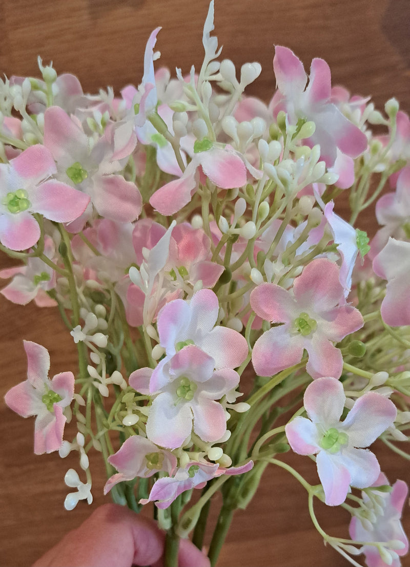 A close-up of a bouquet of artificial hydrangea with baby pink and white flower heads with green stems, held by a hand against a wooden background.