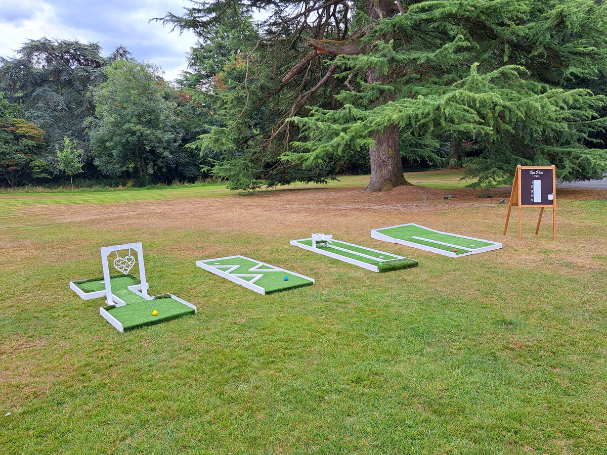 Outdoor mini golf course setup on a grassy lawn with four green wedding themed holes, golf balls, and a blackboard scorecard on an easel, surrounded by trees. Bartle Hall, Preston.