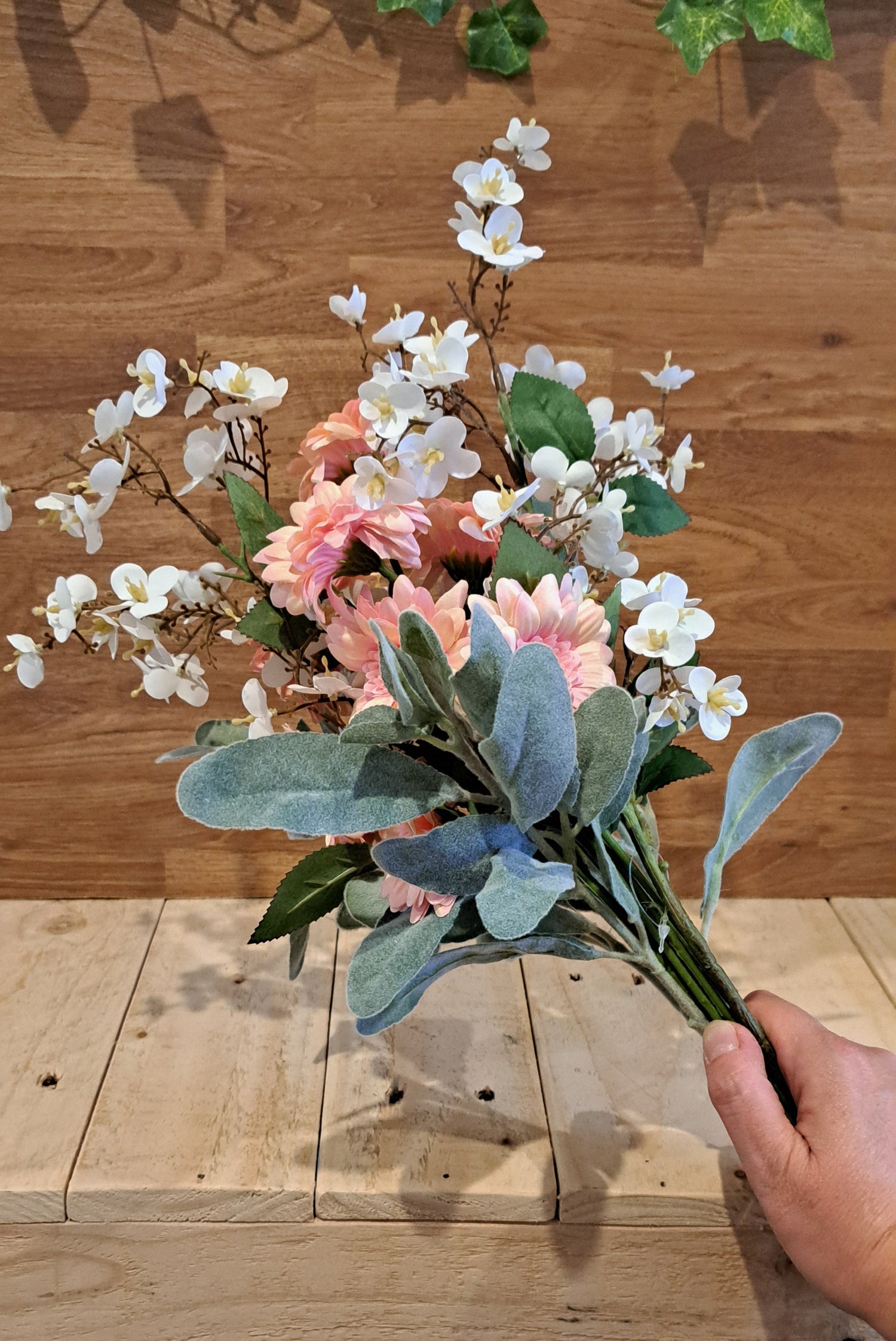 A hand holding a faux floral arrangement with white blossoms, pink flowers, and green and silver-gray leaves, set against a wooden wall and bench.
