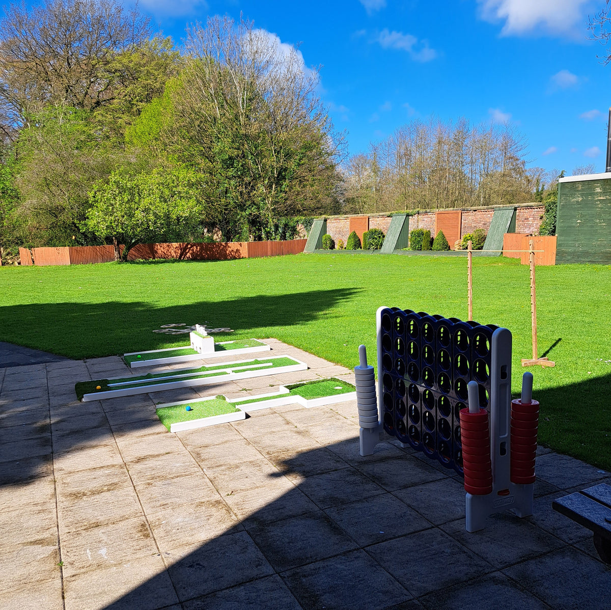 3 holes of wedding mini golf with a giant connect 4 game & limbo setup on a stone patio with a green grass field, trees, blue sky at Briars Hall Hotel in Burscough.