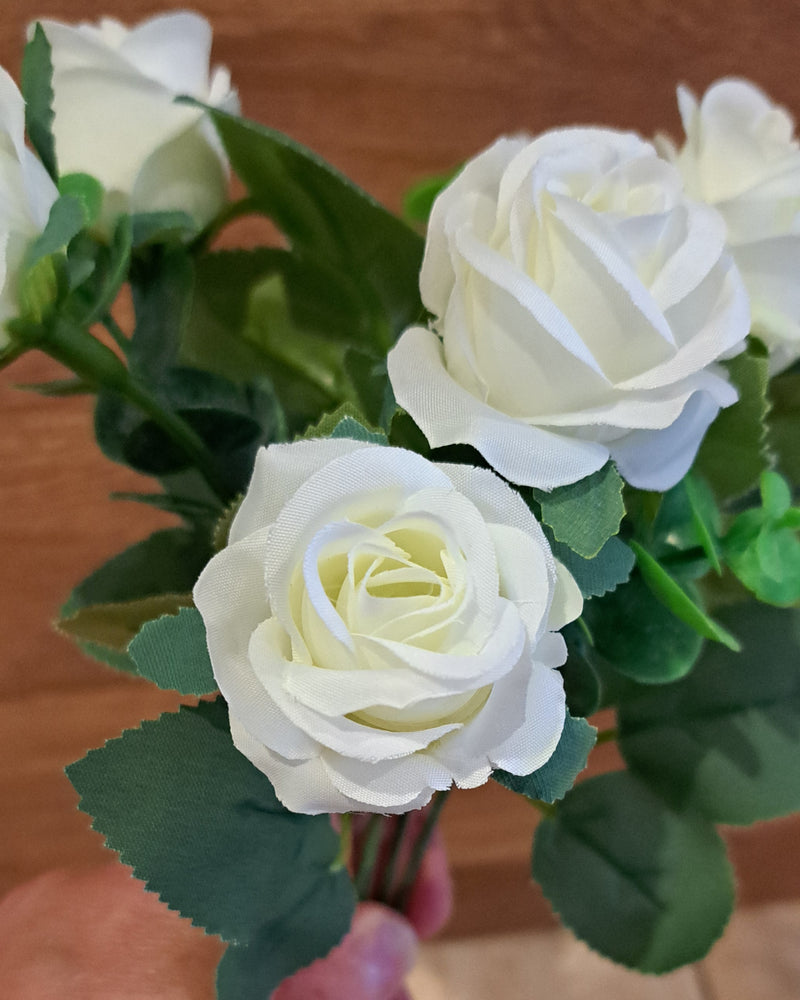 White faux rose artificial flower bouquet with green leaves, held by a hand against a wooden background.