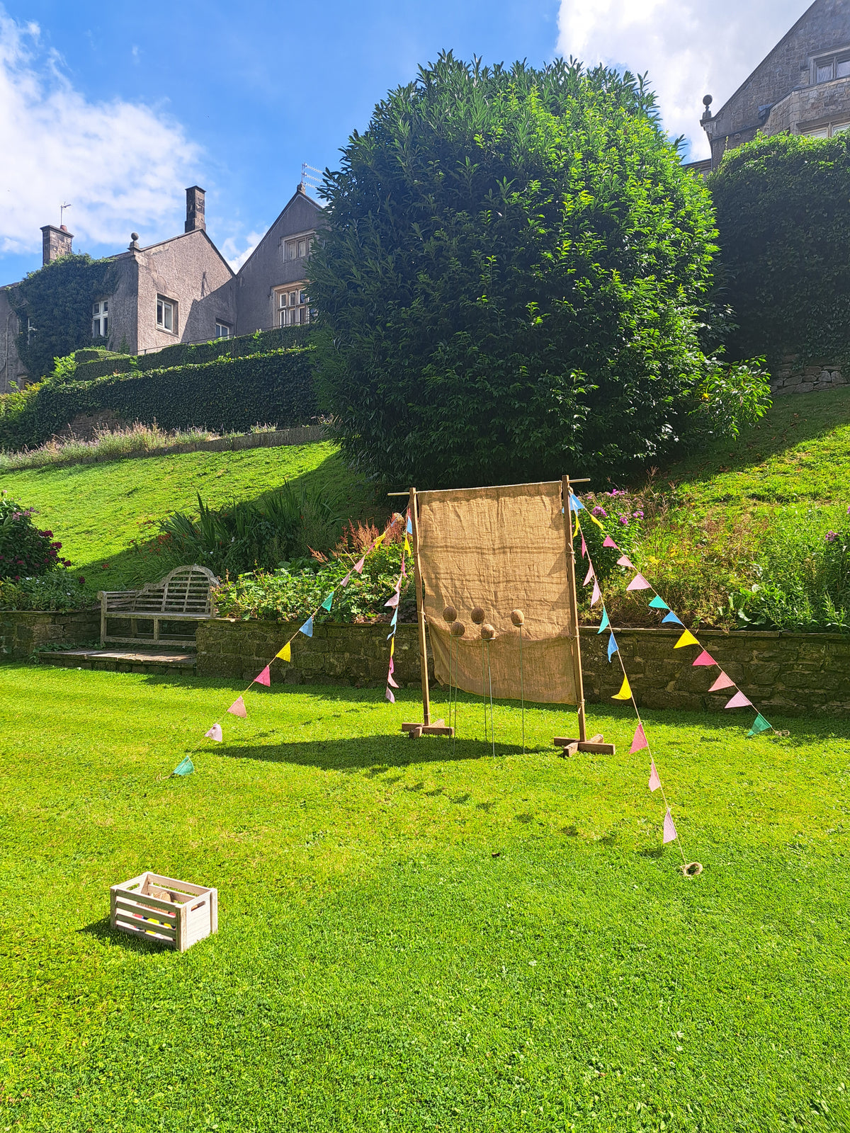 A rustic coconut shy game with burlap covered backdrop set up on a green lawn outdoors, with colourful bunting flags strung between the posts. A house visible in the background under a blue sky. Mitton Hall, Clitheroe.