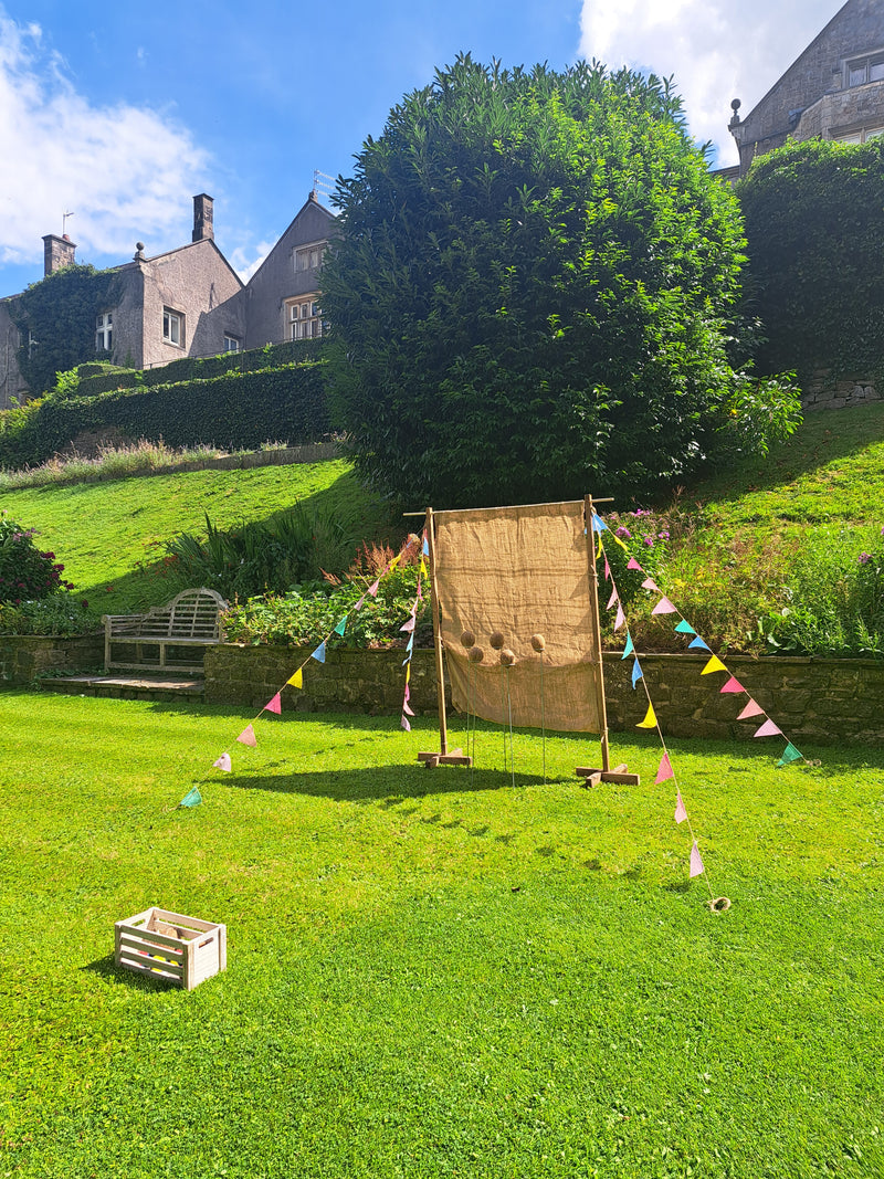 A rustic coconut shy game with burlap covered backdrop set up on a green lawn outdoors, with colourful bunting flags strung between the posts. A house visible in the background under a blue sky. Mitton Hall, Clitheroe.