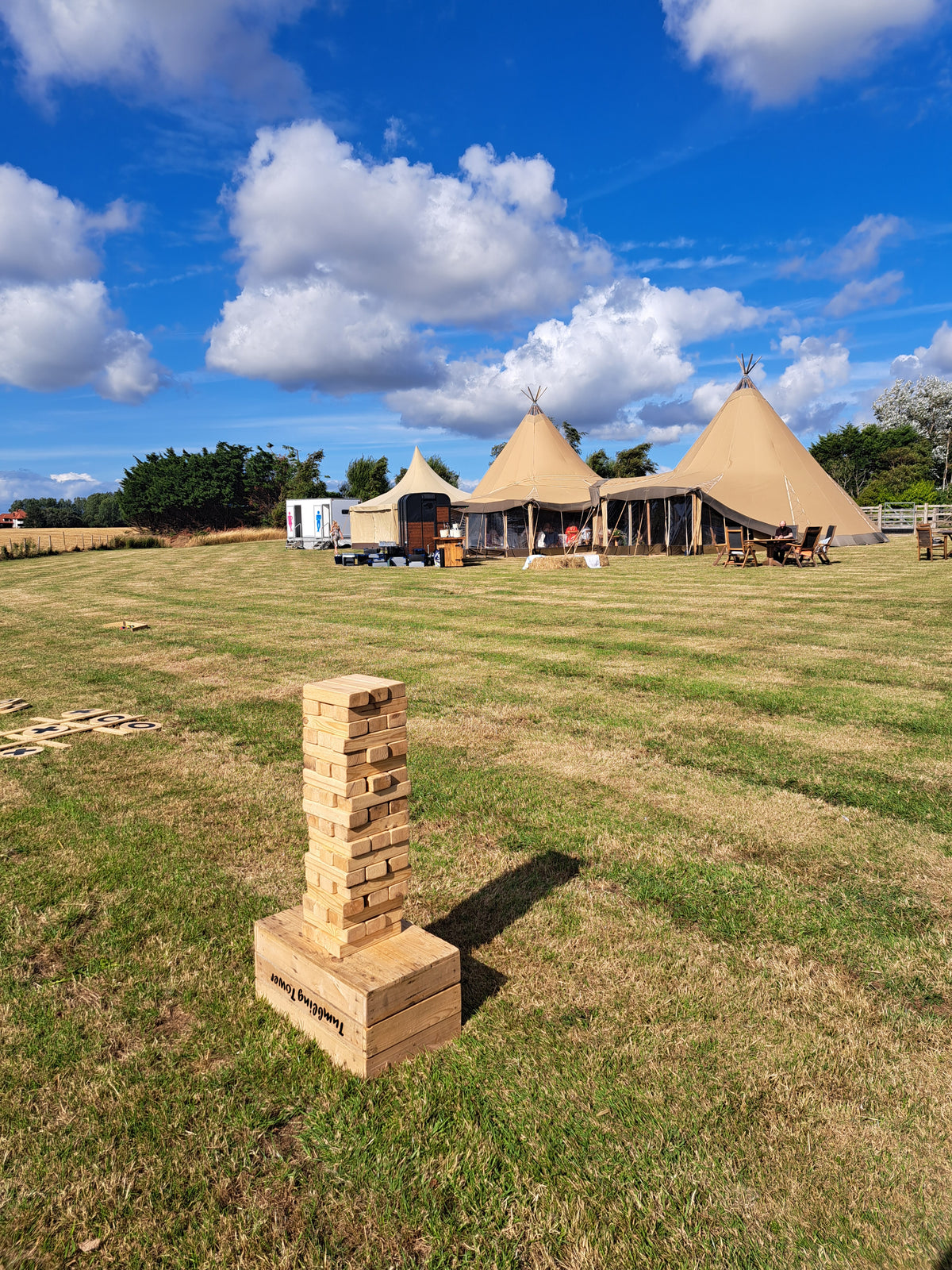 A giant Jenga style wooden block stacking game set up on a grassy field in the foreground, with canvas tipi tents and a blue sky with clouds in the background. Wedding at a private residence, Poulton-le-Fylde.