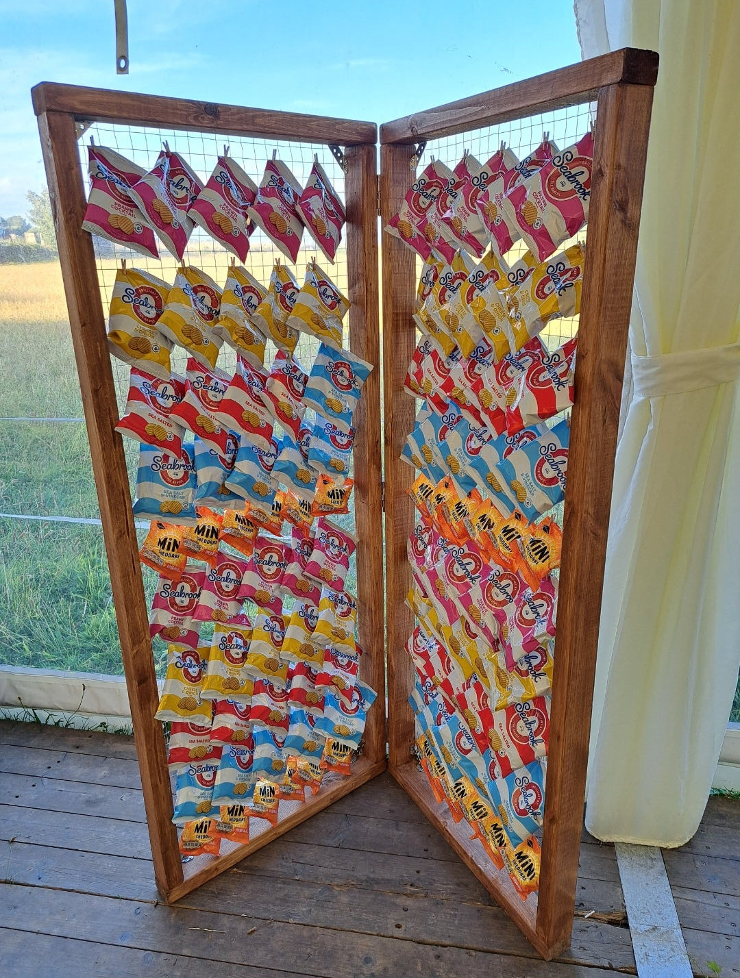 A freestanding crisp wall made up of a folding screen made of wood frames and covered with packets of crisps, on a wooden floor in a marquee with a clear window behind, through which a field and blue sky in the background can be seen.